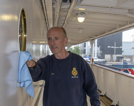 A man polishing the outside on a porthole on the Shelter Deck. 