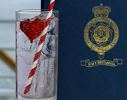 A pink gin fizz in a tall glass with a stripy straw and strawberry next to a navy blue Britannia Tearoom menu. 