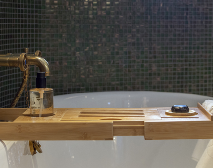 A wooden bath tray sitting atop a bath in Skerryvore Suite's bathroom with body wash, soap and washcloths. The wall is green mosaic tiles in the background. 