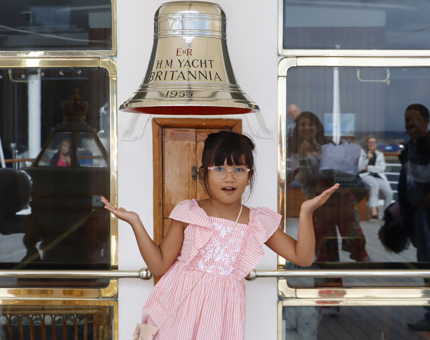 A young girl wearing a pink dress poses in front of The Royal Yacht Britannia's Bell. 