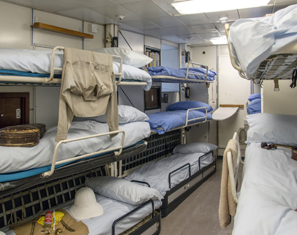 The Royal Marines' Barracks with bunk beds. Musical instruments and uniforms lie on some of the beds. 