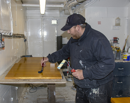 A Maintenance man paints varnish onto a wooden desk in the workshop.