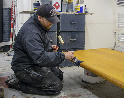 A man from the Maintenance team varnishes the top of the wooden desk in the workshop. 