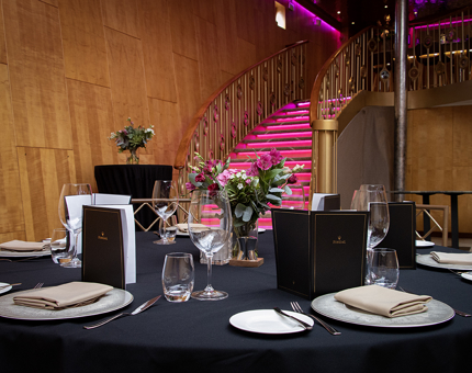 A table set for a private event on Fingal with cutlery, glasses, flowers and menus. The sweeping staircase in the background is illuminated in pink. 