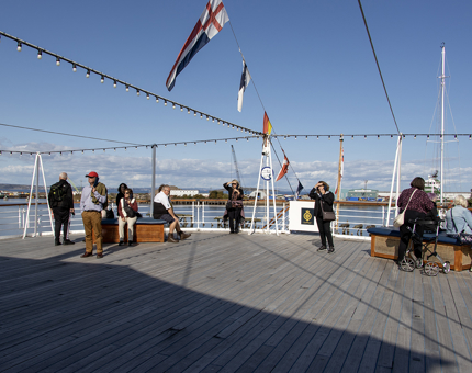 Several visitors standing on the Verandah Deck listening to audio guided tour handsets. 