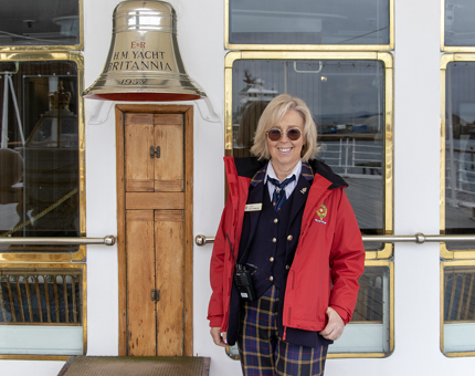 A female Visitor Assistant wearing a Royal Yacht Britannia uniform, smiling and wearing sunglasses, stands next to Britannia's Bell. 