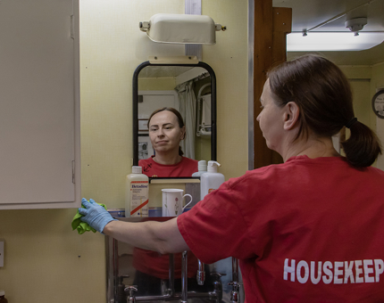 A Housekeeper dusting a tray in the Sick Bay. Her reflection is in the mirror on the wall. 