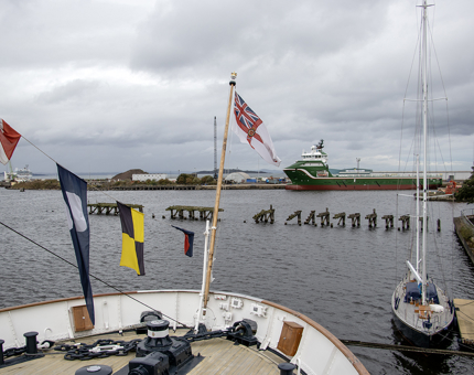 The aft of Britannia with multi-coloured flags flying and a view over the water at Leith Docks with an abandoned pier. 