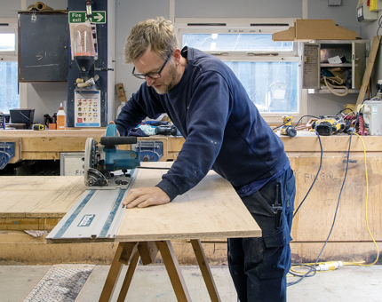 A member of the Maintenance Team using a cutting machine to create flooring for size in the workshop.