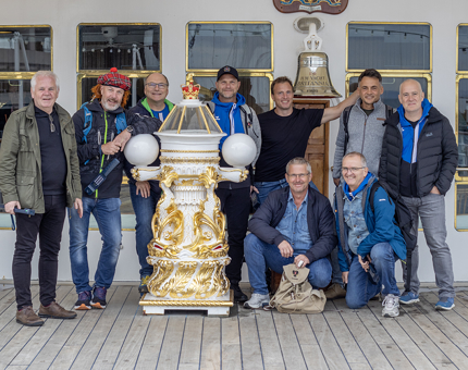 A group of nine men pose standing next to Britannia's binnacle.