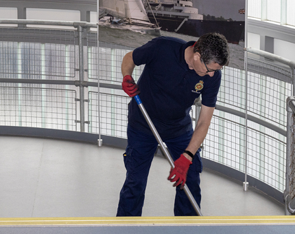 A man sweeping with a broom in the visitor lift tower. A photo of Britannia is hanging on the wall in the background. 