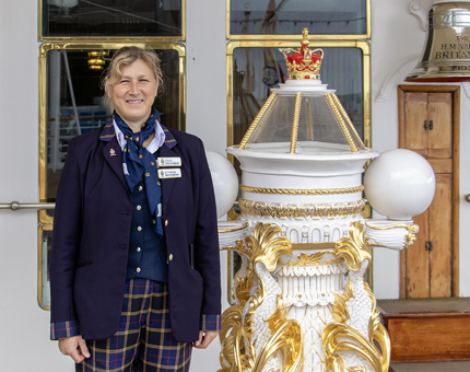 A woman Visitor Assistant stands smiling in uniform at the compass on the Verandah Deck. 