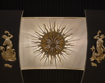 Gold detail on a fireplace with a sun motif and two mythological figures either side.