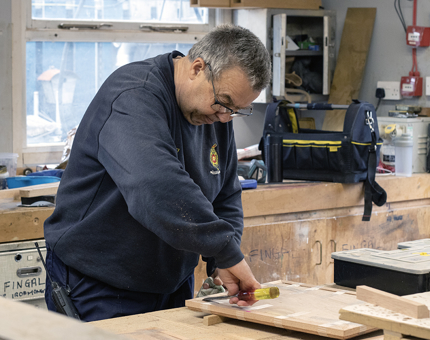 A member of the Maintenance team uses a chisel to build a wooden lectern. 