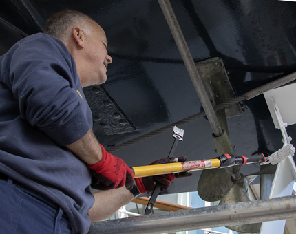 A man from the Maintenance team using a roller to paint underneath a lifeboat.