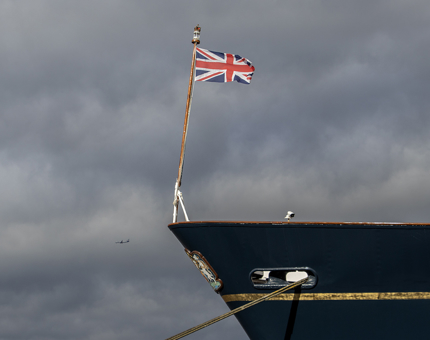 The bow of The Royal Yacht Britannia. The Union Jack flag is flying and an aeroplane is flying past in the background. 