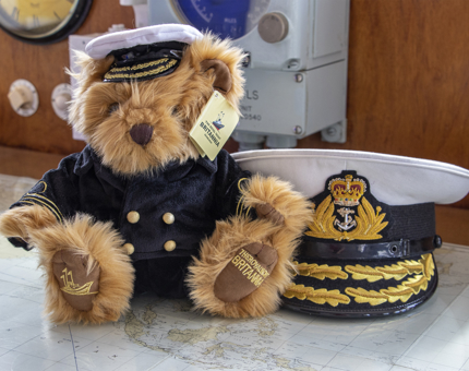 A soft toy teddy bear wearing a Captain's uniform next to a novelty Captain's hat. 