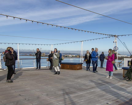 A wide view of the Verandah Deck on a sunny day. Several visitors are listening to the audio guide handsets. 