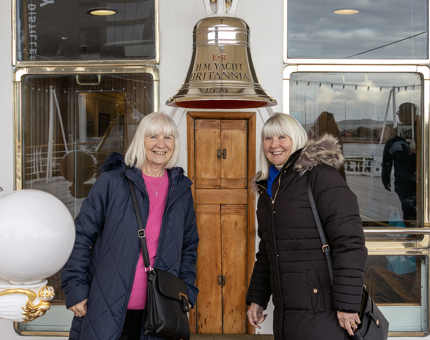 Two women standing next to The Royal Yacht Britannia's bell on the Verandah Deck. 