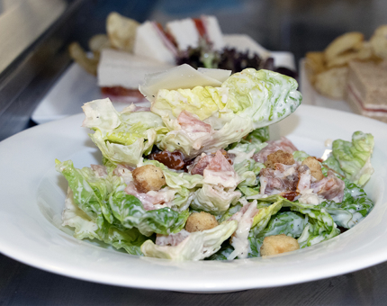 A Caesar salad on a plate with sandwich platters in the background.