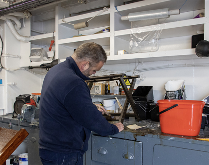 A man from the Maintenance Team varnishing a suitcase luggage rack in the workshop.