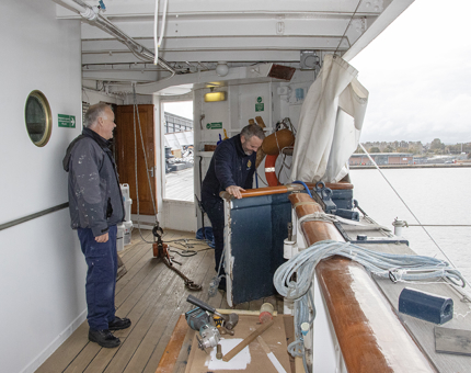 The two men open a door on the side of Britannia. There are tools and ropes on the deck.