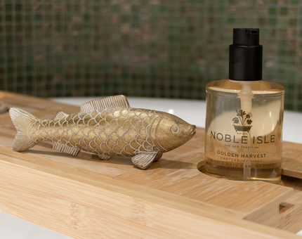 A bottle of Noble Isle shower gel on a bath tray. An ornamental gold fish is on the tray too. 