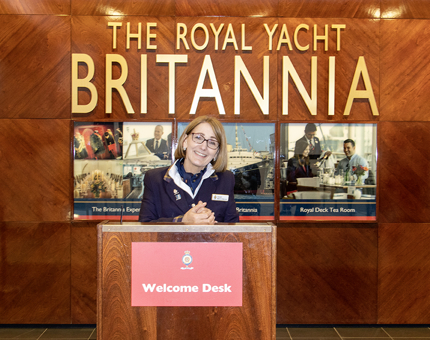A Welcome Host standing smiling at the podium at The Royal Yacht Britannia's Reception. 