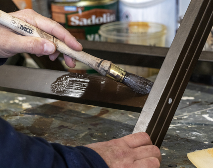 A close-up of a man painting varnish onto a dark brown wooden luggage rack.