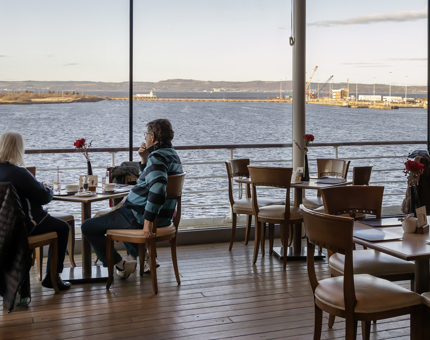 Several tables of visitors in the Royal Deck Tearoom. The sea can be seen outside the windows. 