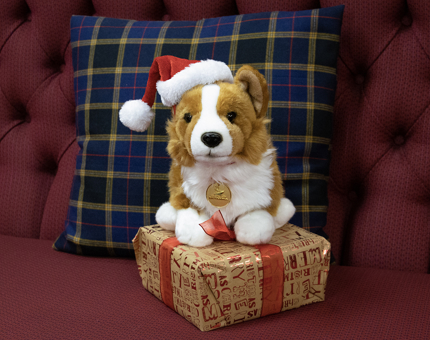 A cuddly Corgi toy sitting on a box wrapped with Christmas paper. The toy is wearing a Santa hat. 