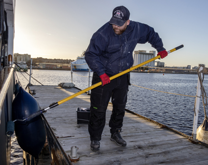 A man using a long roller to paint the outside of Britannia. He is standing on a floating platform. 