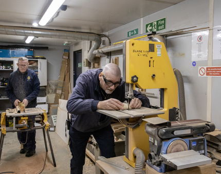 Two men working in the Workshop. One is using a circular saw and the other is cutting a wooden bung. 
