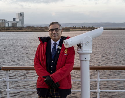 A member of the Visitor Experience team standing on the outside Verandah Deck next to a telescope. The water is in the background. 