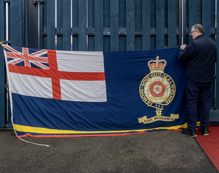 A member of the Maintenance team holding a large flag that contains the Union Jack, St George Cross and Royal Yacht Britannia crest. 