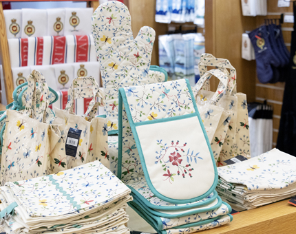 Bags, tea towels, oven gloves and oven gauntlets arranged in a display in the Gift Shop. They all are decorated with a pretty floral design. 