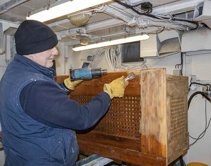 A man using a heatgun to remove varnish from a wooden rope box in the Maintenance workshop.
