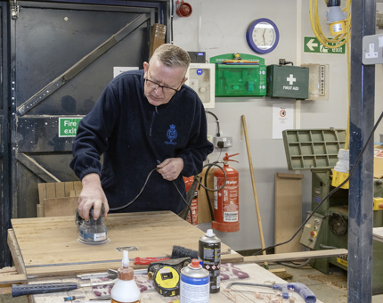 A member of the Maintenance team sanding wood in the workshop. 