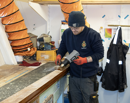A man from the Maintenance team using a heat gun in the workshop to remove varnish from a wooden handrail.