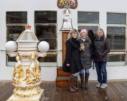 Three women wearing coats and holding a small toy dog pose next to Britannia's bell. 
