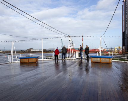 Two couples listen to audio guide handsets while walking on the Verandah Deck. 