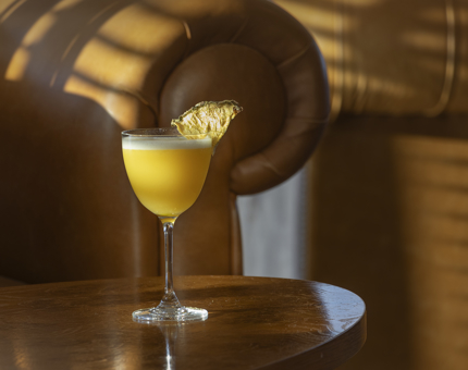 A yellow-coloured mocktail in a wine glass with a slice of pineapple on the side. A tan-coloured leather armchair is in the background.