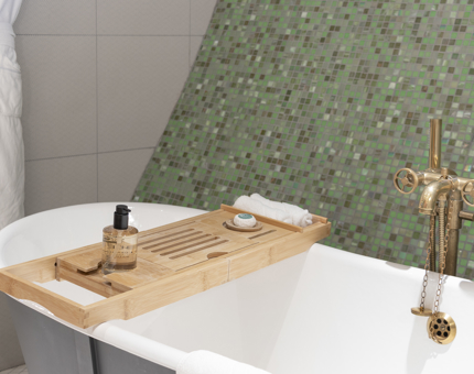 A free-standing bathtub with wooden bath tray and green mosaic wall.