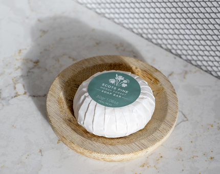 A wrapped bar of Noble Isle Scots Pine soap in a wooden soap dish. 