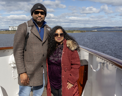 A man and a woman standing on The Royal Yacht Britannia's Bridge. It is a sunny day and they are wearing sunglasses.