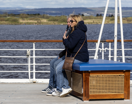 A couple sit on a rope box on the outside Verandah Deck while listening to the audio guided tour.