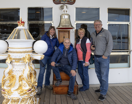 Two men and two women standing in front of The Royal Yacht Britannia's Bell and next to the compass binnacle. 