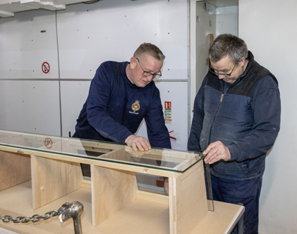 Two men in the Maintenance workshop are assembling a new wooden disaply counter. 