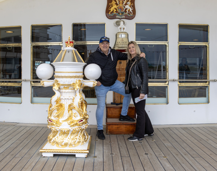 A man and a woman standing next to The Royal Yacht Britannia's Bell on the Verandah Deck. 