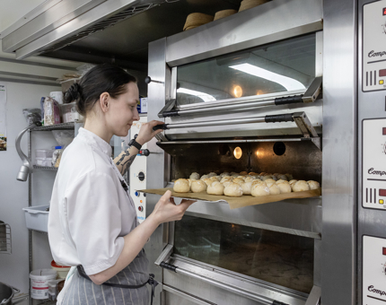 A Chef taking a tray of bread rolls out of an oven in the Galley. 